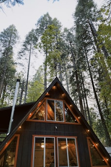 An A-frame cabin is nestled among tall pine trees. The cabin has large triangular windows that allow a glimpse of its warmly lit interior. Strings of lights decorate the cabin's exterior, adding a cozy ambiance.