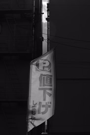 A black and white photograph of an urban setting featuring a vertical banner with Japanese text. The banner is prominently placed in the foreground, while the background shows the outlines of buildings and a fire escape staircase. The lighting suggests a subdued and slightly dramatic atmosphere.