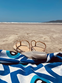 A pair of flip-flops rests on a vibrant blue and white patterned beach towel. The scene is set on a sandy beach, with the ocean in the distance and a clear blue sky overhead.