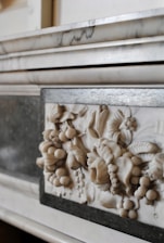 A close-up of a marble relief with intricate carvings of fruits and floral patterns, set within a polished stone frame. The relief showcases detailed leaves, grapes, and other botanical elements in a classic style.