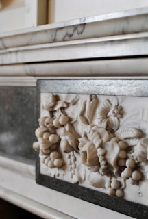 A close-up of a marble relief with intricate carvings of fruits and floral patterns, set within a polished stone frame. The relief showcases detailed leaves, grapes, and other botanical elements in a classic style.