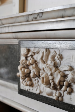 A close-up of a marble relief with intricate carvings of fruits and floral patterns, set within a polished stone frame. The relief showcases detailed leaves, grapes, and other botanical elements in a classic style.