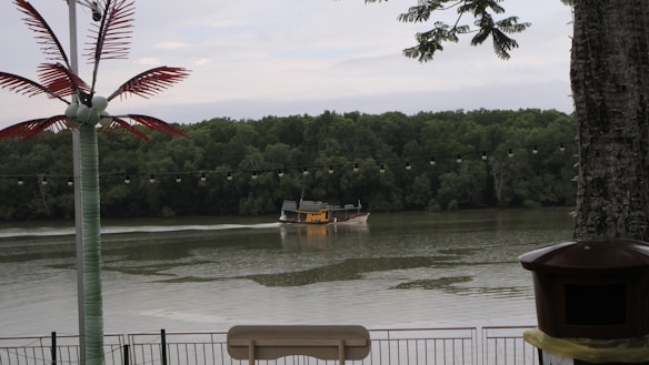 A boat is travelling slowly on a wide river, surrounded by lush green trees on the opposite bank. In the foreground, there is a decorative palm tree made of synthetic material and a string of round lights hanging across the scene. Part of a railing and what appears to be an outdoor furniture or a signpost are visible along with a tree trunk on the right edge.