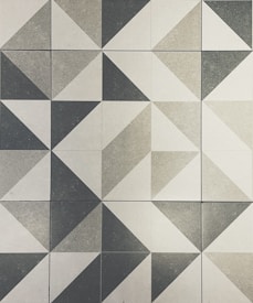 A geometric pattern composed of square tiles arranged in a grid. Each tile contains two triangles that create a variety of shapes, with some resembling parallelograms and others forming larger diamond shapes. The tiles are in shades of gray, with subtle variations creating a textured appearance.
