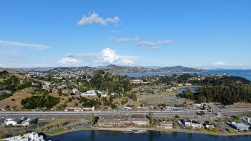 Aerial view of a coastal area with residential neighborhoods and greenery. Hills and mountains are visible in the background, leading to a body of water that meets the sky at the horizon. Roads and buildings are present in the foreground, with a mix of white clouds and blue sky above.