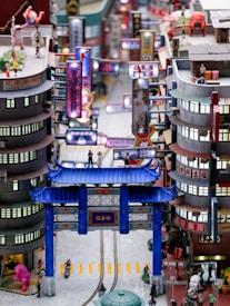 A detailed miniature cityscape features brightly colored signage with Chinese characters, people and robots in various poses, and a prominent blue traditional Chinese gate. Buildings are closely packed with intricate details, and small figures engage in different activities. The scene is lively with a blend of modern and traditional elements.