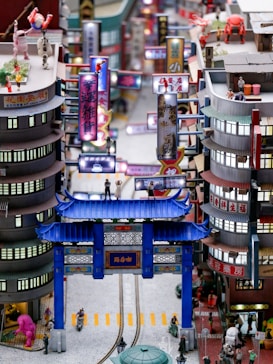 A detailed miniature cityscape features brightly colored signage with Chinese characters, people and robots in various poses, and a prominent blue traditional Chinese gate. Buildings are closely packed with intricate details, and small figures engage in different activities. The scene is lively with a blend of modern and traditional elements.