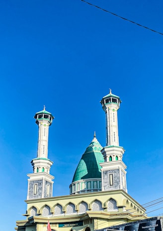 A mosque with two tall minarets featuring intricate architectural details and a large central dome with a green top. The structure is set against a clear blue sky, with some power lines visible in the foreground.