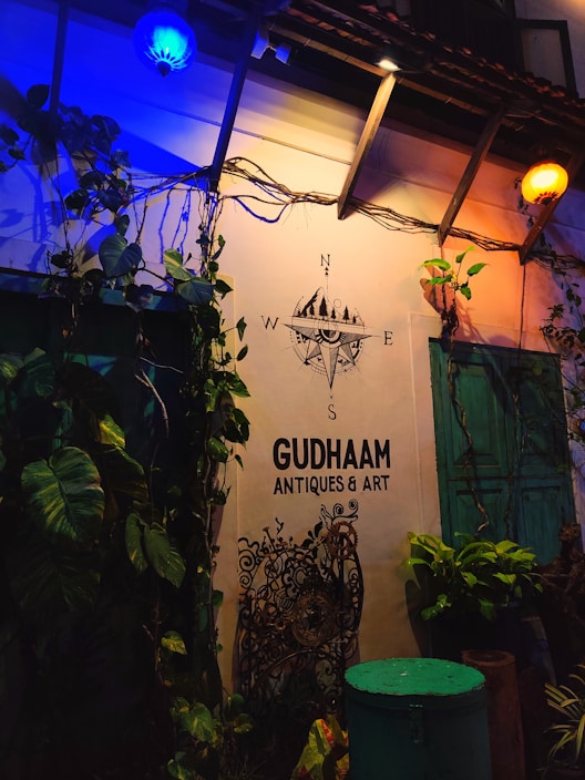The scene depicts an exterior wall of an establishment named GUDHAAM ANTIQUES & ART. Illuminated by both blue and yellow lights, the wall is adorned with green leafy plants and vintage decorations. A nautical compass decal with cardinal directions is displayed above the business name. There is a turquoise-colored door and some potted plants below.