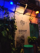 The scene depicts an exterior wall of an establishment named GUDHAAM ANTIQUES & ART. Illuminated by both blue and yellow lights, the wall is adorned with green leafy plants and vintage decorations. A nautical compass decal with cardinal directions is displayed above the business name. There is a turquoise-colored door and some potted plants below.