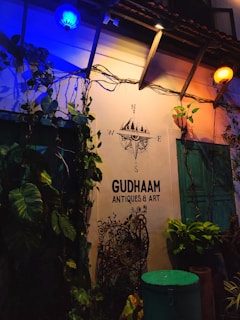 The scene depicts an exterior wall of an establishment named GUDHAAM ANTIQUES & ART. Illuminated by both blue and yellow lights, the wall is adorned with green leafy plants and vintage decorations. A nautical compass decal with cardinal directions is displayed above the business name. There is a turquoise-colored door and some potted plants below.