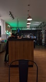 A modern cafe setting with a wooden counter surrounded by people in a dimly lit environment. Pendant lights hang overhead, casting soft light. A neon sign in the background contrasts with the warm wooden floors.