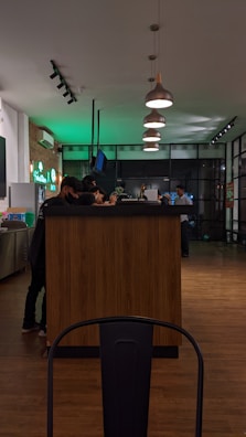 A modern cafe setting with a wooden counter surrounded by people in a dimly lit environment. Pendant lights hang overhead, casting soft light. A neon sign in the background contrasts with the warm wooden floors.