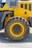 A close-up view of a large, rugged tire attached to a piece of heavy machinery, likely construction equipment. The body of the vehicle is painted bright yellow with some gray elements and appears to be on a gravel surface, suggesting an outdoor, industrial setting.