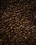 A close-up view of a large pile of roasted coffee beans, showcasing their rich and varied brown tones. The beans are densely packed, with some surface sheen indicating freshness.