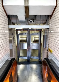 A narrow corridor featuring a turnstile entrance commonly found in subway or metro stations. The walls are lined with white subway tiles, and two metallic turnstiles stand at the front. An escalator handrail is visible leading towards the turnstile, indicating an entry point for passengers.