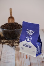 A package of Sleepy Owl Original Coffee is prominently displayed in the foreground. The package is colored in blue and white and is labeled as Pure Arabica. Behind the package, there is a glass bowl filled with coffee beans, with a wooden scoop placed upright in the bowl. The scene is set on a wooden surface, enhancing the rustic and cozy atmosphere.
