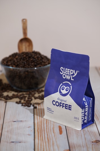 A package of Sleepy Owl Original Coffee is prominently displayed in the foreground. The package is colored in blue and white and is labeled as Pure Arabica. Behind the package, there is a glass bowl filled with coffee beans, with a wooden scoop placed upright in the bowl. The scene is set on a wooden surface, enhancing the rustic and cozy atmosphere.