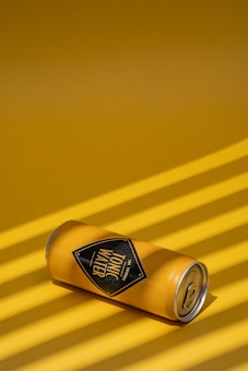 A can of tonic water is laying on its side against a solid yellow background. The surface of the can appears to be cold, with condensation droplets visible. Shadows created by parallel lines add texture and depth to the image.