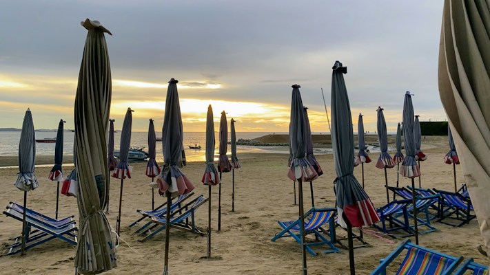 A serene beach scene with numerous closed beach umbrellas and striped lounge chairs scattered across the sandy shore. They are facing towards the ocean where a gentle sunset paints the horizon in shades of orange and yellow. The sky is partially cloudy, adding a tranquil and peaceful atmosphere to the setting.