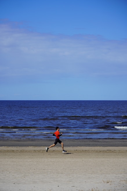 A person wearing a red shirt and black shorts is jogging along a sandy beach with the ocean in the background. The sky is mostly clear with a few clouds, and the waves are gently lapping at the shore.