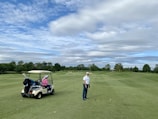 A person wearing a cap and holding a golf club stands on a well-maintained golf course. A golf cart nearby has a passenger wearing a pink jacket and a large white hat. The background features lush green trees and a partly cloudy sky.
