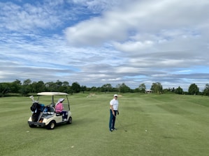 A person wearing a cap and holding a golf club stands on a well-maintained golf course. A golf cart nearby has a passenger wearing a pink jacket and a large white hat. The background features lush green trees and a partly cloudy sky.