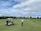 A person wearing a cap and holding a golf club stands on a well-maintained golf course. A golf cart nearby has a passenger wearing a pink jacket and a large white hat. The background features lush green trees and a partly cloudy sky.