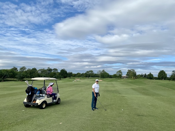 A person wearing a cap and holding a golf club stands on a well-maintained golf course. A golf cart nearby has a passenger wearing a pink jacket and a large white hat. The background features lush green trees and a partly cloudy sky.