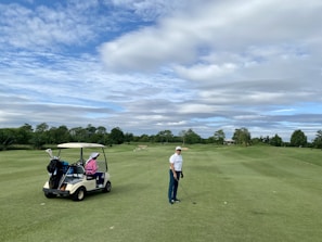 A person wearing a cap and holding a golf club stands on a well-maintained golf course. A golf cart nearby has a passenger wearing a pink jacket and a large white hat. The background features lush green trees and a partly cloudy sky.