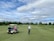 A person wearing a cap and holding a golf club stands on a well-maintained golf course. A golf cart nearby has a passenger wearing a pink jacket and a large white hat. The background features lush green trees and a partly cloudy sky.