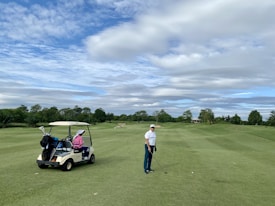 A person wearing a cap and holding a golf club stands on a well-maintained golf course. A golf cart nearby has a passenger wearing a pink jacket and a large white hat. The background features lush green trees and a partly cloudy sky.
