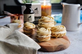 A wooden board holds a small stack of sandwich cookies filled with cream. The cookies are neatly piped and set in an inviting kitchen atmosphere. A bottle labeled 'vanilla' sits in the background, alongside a lit candle, a pitcher, copper measuring cup, and a linen cloth, adding warmth to the setting.