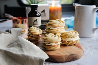A wooden board holds a small stack of sandwich cookies filled with cream. The cookies are neatly piped and set in an inviting kitchen atmosphere. A bottle labeled 'vanilla' sits in the background, alongside a lit candle, a pitcher, copper measuring cup, and a linen cloth, adding warmth to the setting.