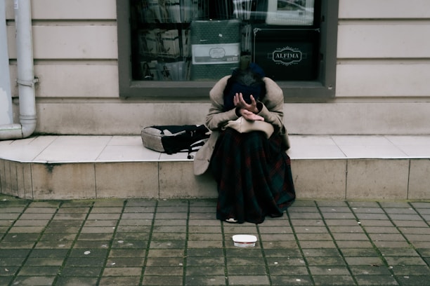 A person is sitting on a sidewalk with their head in their hands, next to a building with a window. Nearby, an open container is placed on the ground, suggesting they might be seeking help or donations. The person is wearing a long coat and a blanket, indicating possible cold weather.