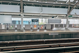 An empty train station platform with signage including a sign for 'Saint Louis' station. The platform features multiple billboards, a safety warning on the edge, and two visible train tracks in the foreground. The surrounding architecture includes large glass windows and industrial-style roofing.