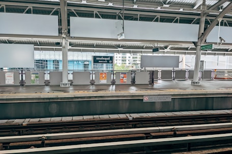 An empty train station platform with signage including a sign for 'Saint Louis' station. The platform features multiple billboards, a safety warning on the edge, and two visible train tracks in the foreground. The surrounding architecture includes large glass windows and industrial-style roofing.