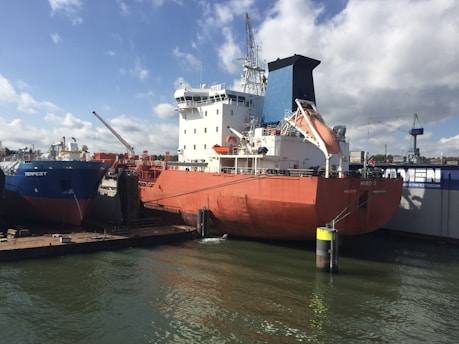 Two large ships docked at a port, one with a blue hull and another with a red hull. The ships are positioned side by side, with cranes and other port equipment visible in the background. A calm body of water surrounds the vessels, and the sky is partly cloudy, allowing sunlight to brightly illuminate the scene.