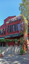 A vibrant red brick building with green shutters and windows. The facade is adorned with hanging purple flowers and green plants, creating a charming and colorful appearance. The establishment appears to be a restaurant with signage reading 'Italian Mutfağı' and 'İtalyan Mutfağı' indicating an Italian cuisine. A tree is visible in the background with a clear blue sky suggesting a bright, sunny day.