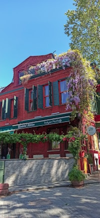 A vibrant red brick building with green shutters and windows. The facade is adorned with hanging purple flowers and green plants, creating a charming and colorful appearance. The establishment appears to be a restaurant with signage reading 'Italian Mutfağı' and 'İtalyan Mutfağı' indicating an Italian cuisine. A tree is visible in the background with a clear blue sky suggesting a bright, sunny day.