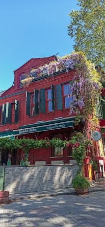 A vibrant red brick building with green shutters and windows. The facade is adorned with hanging purple flowers and green plants, creating a charming and colorful appearance. The establishment appears to be a restaurant with signage reading 'Italian Mutfağı' and 'İtalyan Mutfağı' indicating an Italian cuisine. A tree is visible in the background with a clear blue sky suggesting a bright, sunny day.