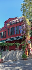 A vibrant red brick building with green shutters and windows. The facade is adorned with hanging purple flowers and green plants, creating a charming and colorful appearance. The establishment appears to be a restaurant with signage reading 'Italian Mutfağı' and 'İtalyan Mutfağı' indicating an Italian cuisine. A tree is visible in the background with a clear blue sky suggesting a bright, sunny day.