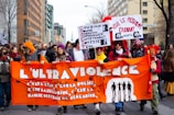 A group of people walking down a street, holding signs and banners protesting various social and political issues. The prominent orange banner at the front reads in French, expressing dissatisfaction with state policies and increased educational fees. The participants are dressed in warm clothing, and some wear colorful hats and masks.