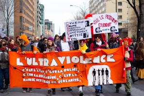 A group of people walking down a street, holding signs and banners protesting various social and political issues. The prominent orange banner at the front reads in French, expressing dissatisfaction with state policies and increased educational fees. The participants are dressed in warm clothing, and some wear colorful hats and masks.