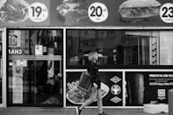 A woman is walking past a storefront with large advertisements for food items featuring prices in Turkish lira. The scene is in black and white, showing the urban environment with a focus on the person carrying a patterned bag. The background includes reflections and signs on the glass windows.