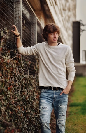 A young adult with curly hair leans casually against a chain-link fence covered in ivy, wearing a white long-sleeve shirt and ripped blue jeans. The setting is an outdoor area with a walkway and a building in the background.