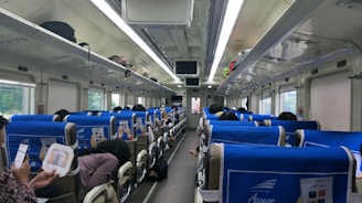 An interior view of a train cabin featuring rows of seats with blue headrest covers. Passengers are seated, some are using their phones, and luggage is stored in the overhead compartments. The ceiling has fluorescent lights and the windows are covered with blinds.
