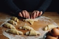 Hands rolling dough into croissant shapes on a floured wooden surface, with several finished croissants nearby and a bowl of eggs in the corner.