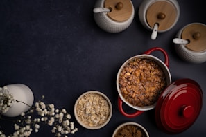 A dark background contrasts with a red casserole dish filled with a baked dish, topped with a crusty brown layer likely made of oats or granola. Surrounding it are neutral-toned ceramic jars with wooden lids, a bowl of oats, a bowl of brown sugar, and a white vase containing small white flowers, creating a rustic yet elegant kitchen scene.