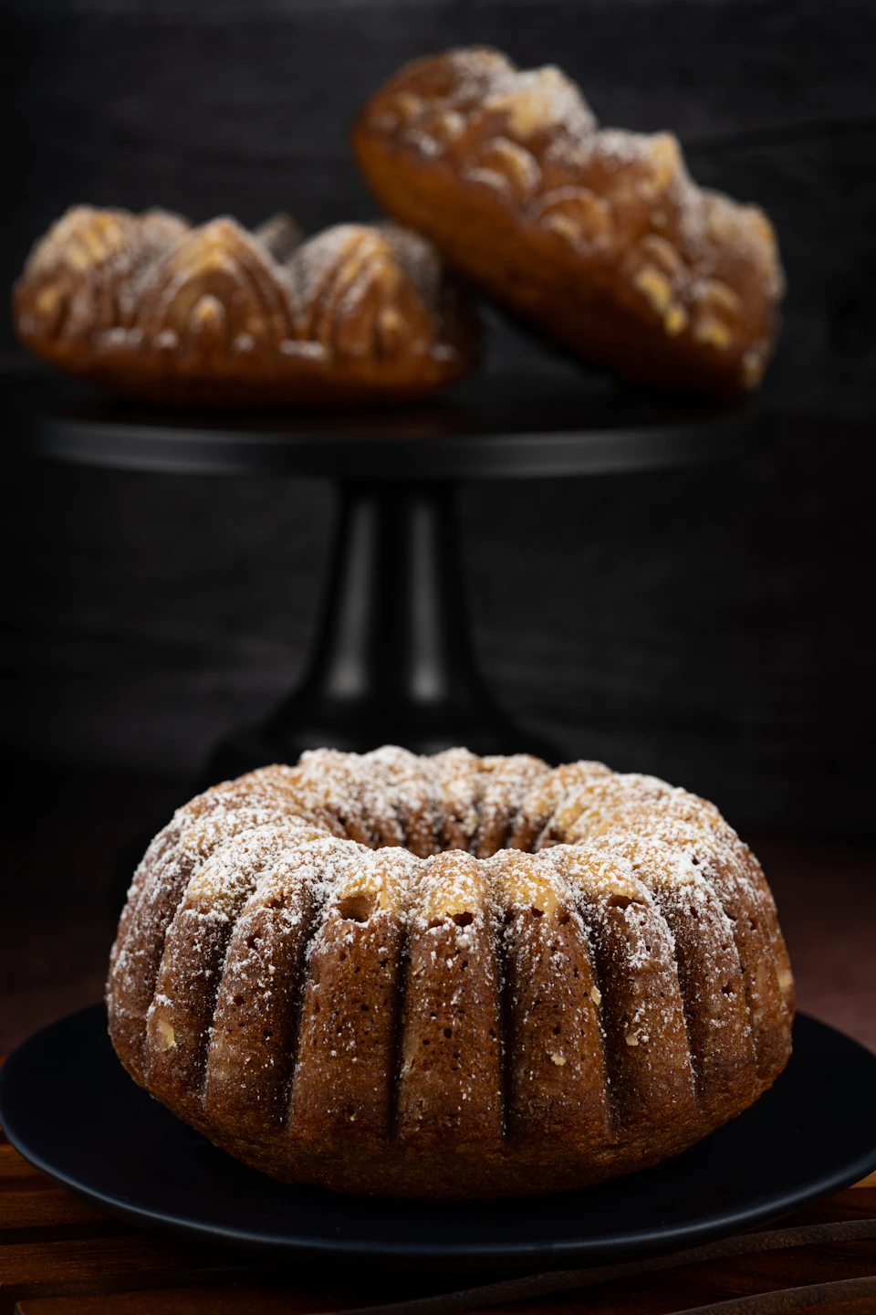 The image presents a delectable dessert scene, featuring three cakes on display against a dark background. The largest cake is placed centrally and is generously covered with powdered sugar, giving it an appetizing appearance. To its left, another cake can be seen, also dusted in powdered sugar, adding to the visual appeal of the desserts. On the right side of the image, there's a smaller cake that shares similar characteristics with the larger one - both are adorned with a generous dusting of powdered sugar.

The cakes are placed on black cake stands, which contrast nicely against the dark background and draw attention to their delicious details. The arrangement of these three cakes creates an enticing visual display, inviting viewers to indulge in this sweet treat.